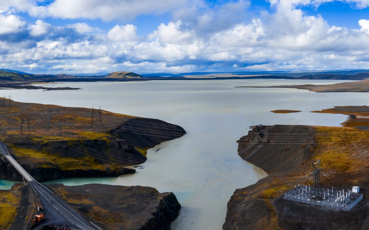 Aerial view shows a milky blue glacial lake near Sigoldugljufur in Iceland. A bridge and road cross a channel, with a substation on lava, clouds diffuse light over basalt.
